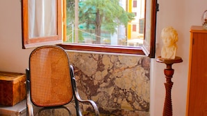 A room with a rocking chair, a bust on a pedestal, and a window with a view of trees and buildings.