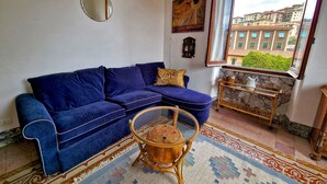 A living room with a blue sofa, a round wooden coffee table, a patterned rug, a window with a view of buildings, and a small side table with a vase.