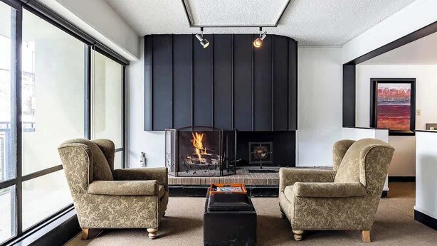 Una sala de estar moderna con chimenea, dos sillones, una mesita de centro y ventanas amplias.