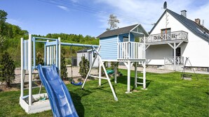 Un terrain de jeux avec un toboggan bleu et des structures d’escalade, une maison blanche dotée d’un balcon et un ciel d’un bleu limpide.