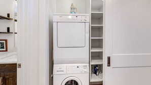 Une petite buanderie avec des appareils superposés, notamment un lave-linge et un sèche-linge, ainsi qu’une petite étagère sur laquelle se trouvent une bouteille et un cadre avec une photo.