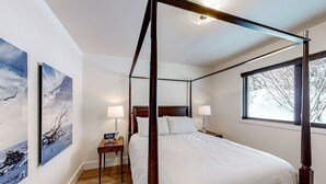 Un lit à baldaquin avec une tête de lit en bois, deux tables de chevet avec des lampes et une grande fenêtre donnant sur les arbres.