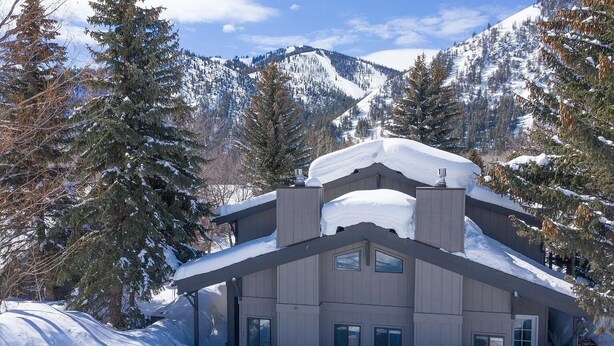 A modern chalet with a symmetrical roof and large windows, surrounded by snow-covered mountains and evergreen trees.