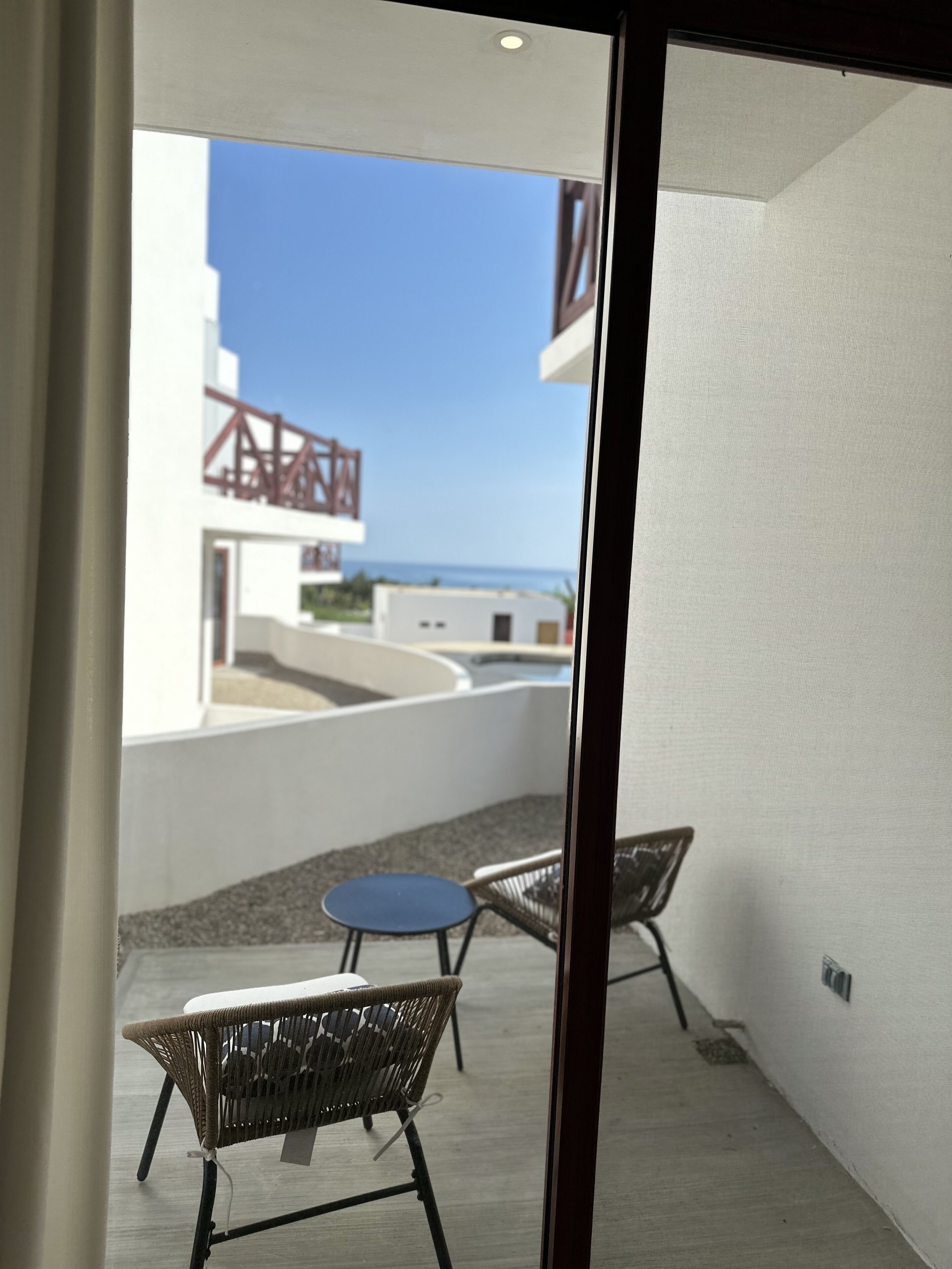 A balcony with wicker chairs and a small table, overlooking a building with a red structure and a view of the sea.