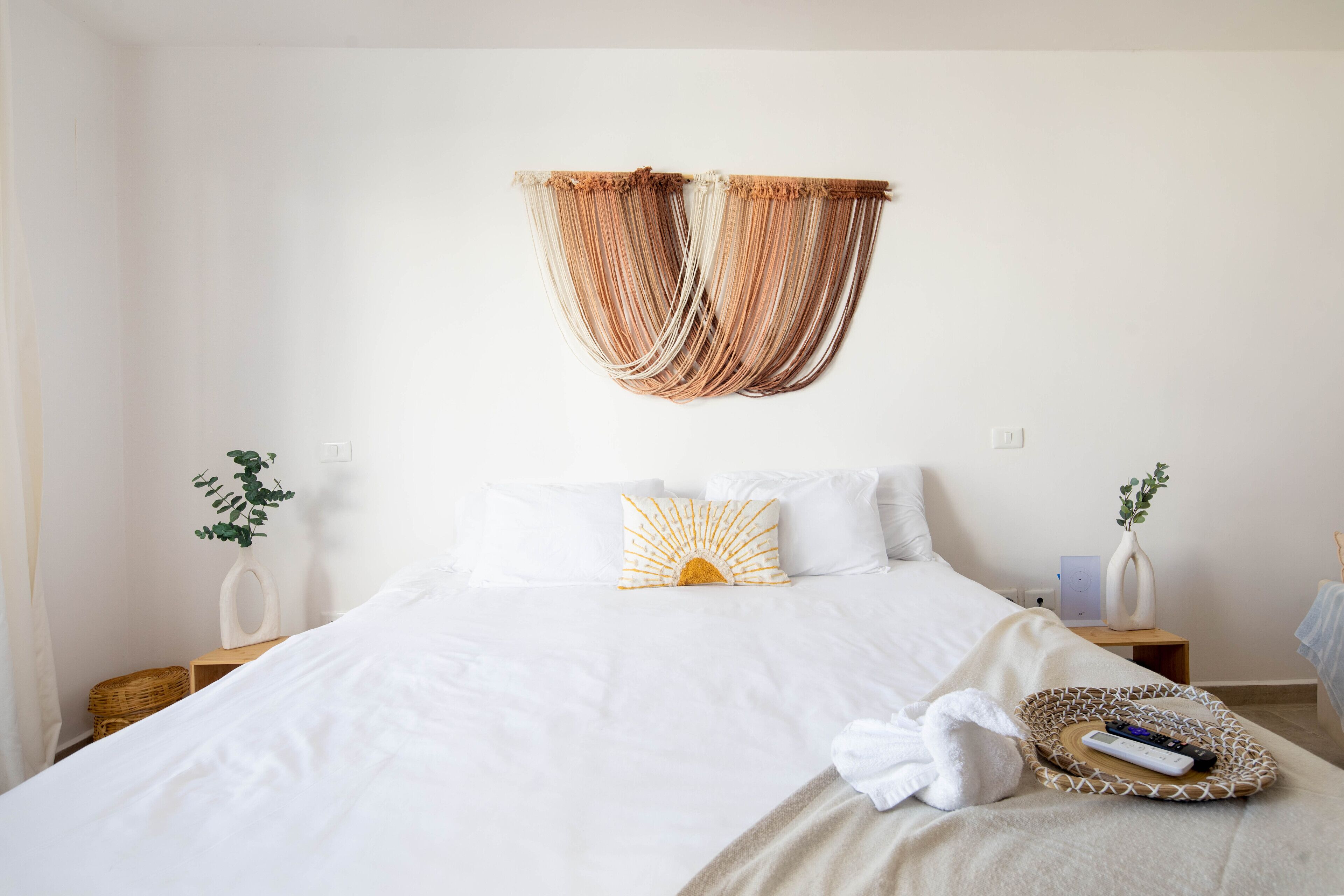 A neatly made bed with white linens, a sunburst pillow, and a remote control on a tray.