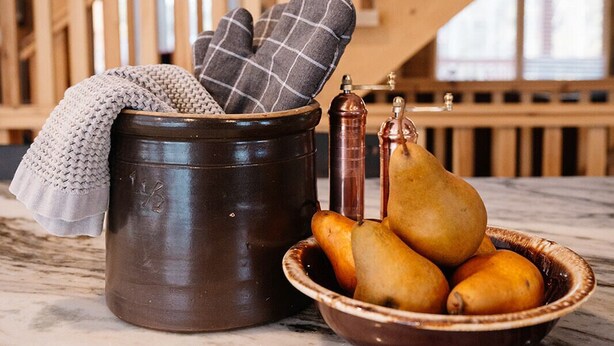 Un ambiente de cocina rústica con una escalera de madera, una olla marrón oscuro, un cuenco de peras y un guante de cocina de cuadros.