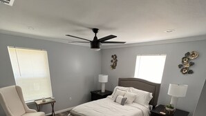A bedroom with a bed, a chair, a nightstand, a lamp, and a ceiling fan.