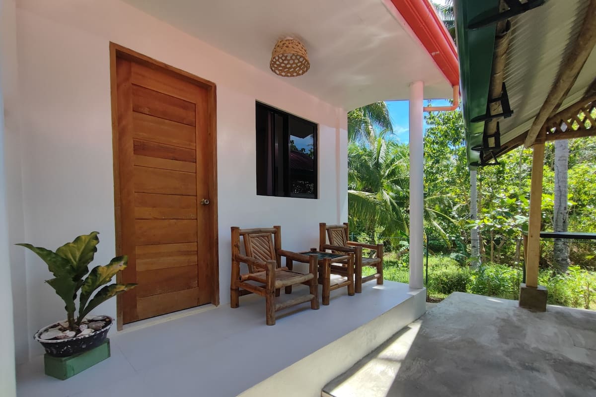 A wooden door, a window, and two wooden chairs on a porch with a view of greenery.