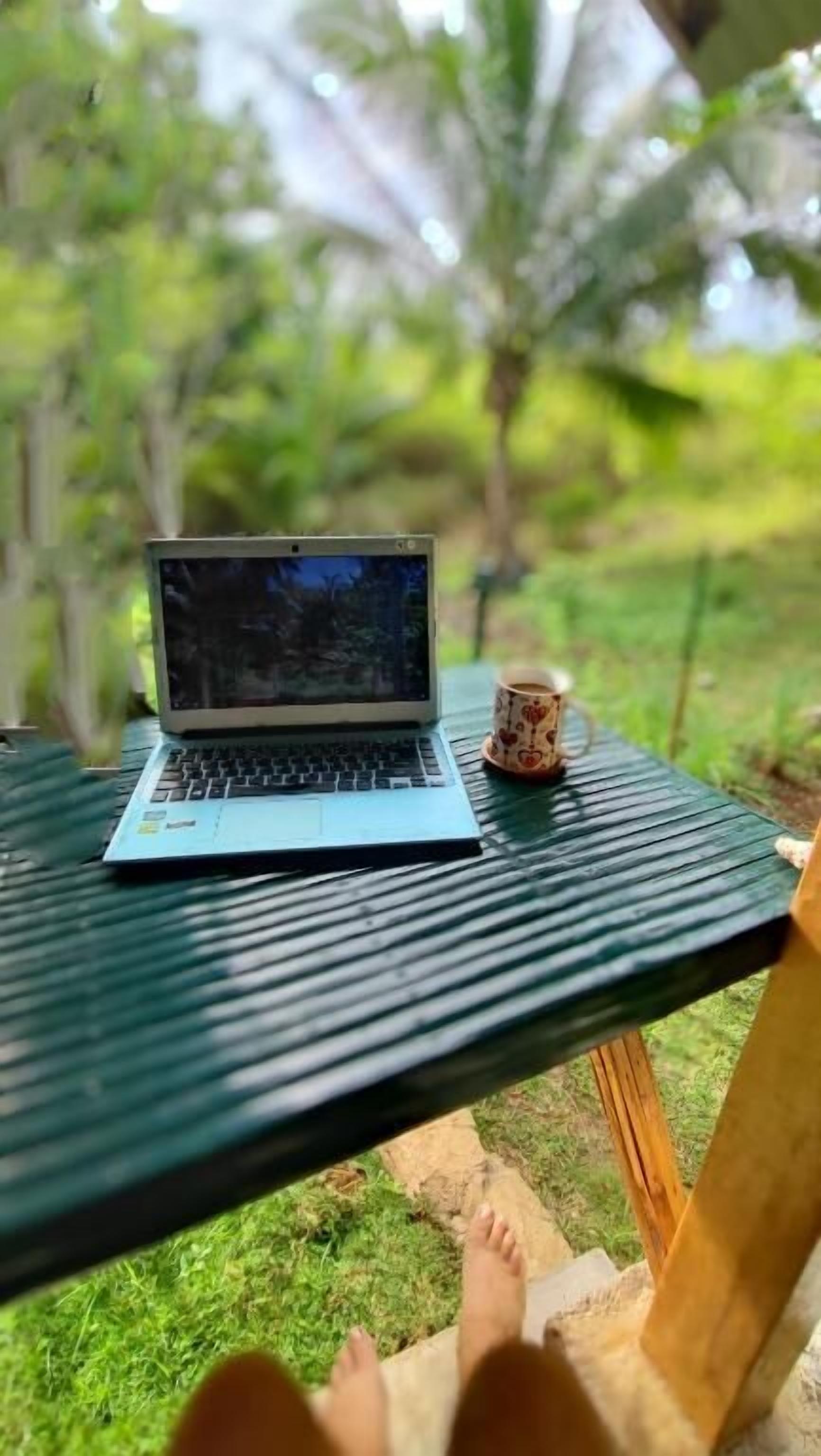 A laptop on a table with a cup beside it, outdoors with greenery in the background.