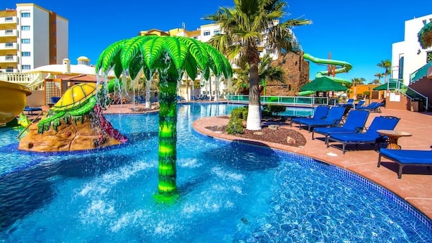 Un área de piscina con una fuente de agua con forma de palmera, sillas de descanso y un tobogán.