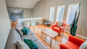 Un vaste espace de vie doté d’un haut plafond, d’un plancher en bois, d’un ensemble de canapés gris, d’un fauteuil rouge et d’une petite table.