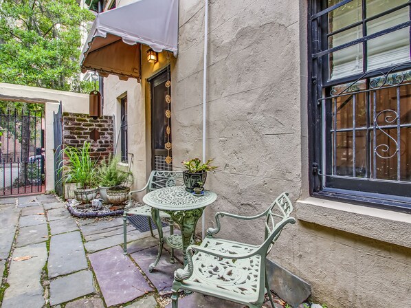 A courtyard with a table and chairs, potted plants, a metal gate, and a building with a textured wall and window with decorative bars. - Enchanted property with a beautiful courtyard! (Savannah)