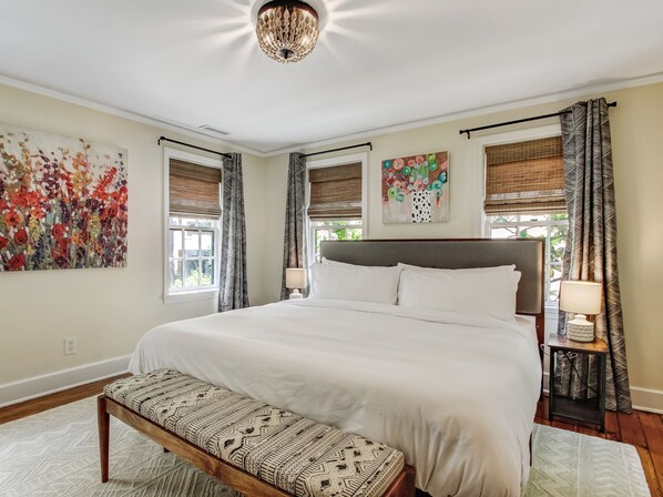 A bedroom with a large bed, a bench, a bedside table with a lamp, and two windows with curtains. - Enchanted property with a beautiful courtyard! (Savannah)