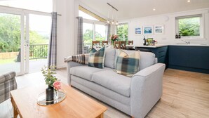 A spacious living area with a grey sofa, wooden coffee table, and a dining table with chairs. A kitchen area with a sink and cabinets is visible in the background. - Eden Fields Lodge (Cupar)