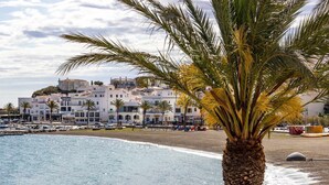 A coastal building with multiple floors, a beach in the foreground, and palm trees. - Port de Llançà 29 – Apartment with sea views (Llança)
