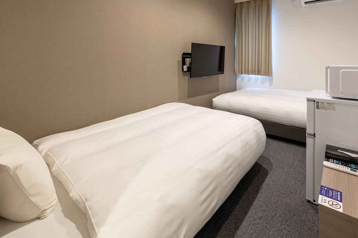 A hotel room with two single beds, a flat-screen TV mounted on the wall, a minibar, and an air conditioning unit.