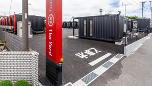 Entrada de hotel com placa vermelha, parede de tijolos e área de estacionamento com cerca.