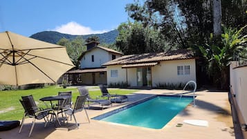 Zona de piscina con sillones reclinables y un gran paraguas, una casa con techo de tejas y una montaña al fondo.