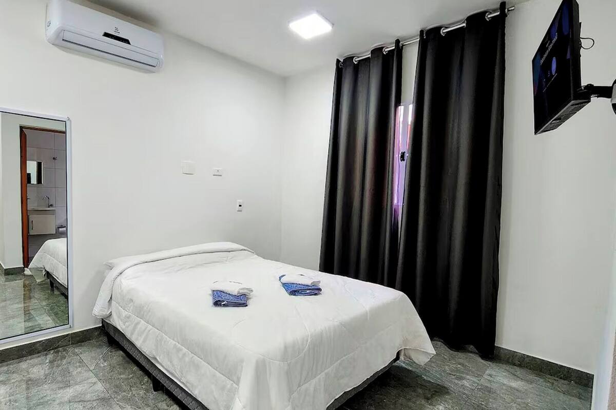 A small, clean room with a single bed, a mirror, an air conditioning unit, and a TV mounted on the wall.