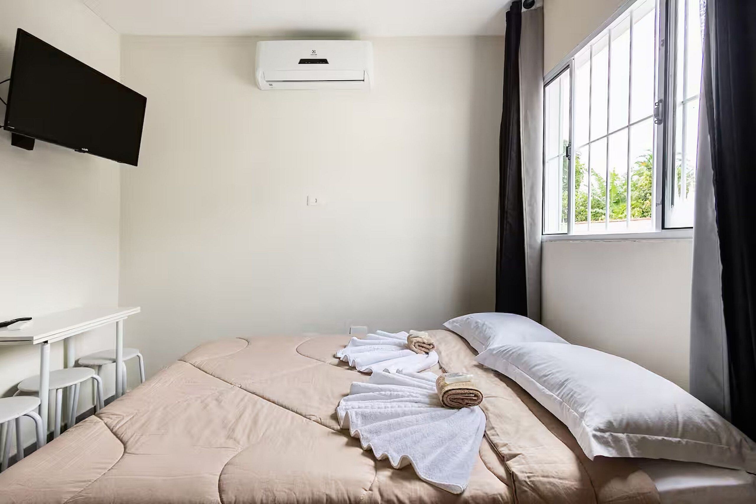 A hotel room with a bed, a desk, a flat-screen TV mounted on the wall, and a window with curtains.
