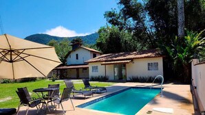 A pool area with lounge chairs, a large umbrella, and a house with a tiled roof. - Maresias Suítes (São Sebastião)