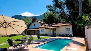 Zona de piscina amb gandules, un parasol gran i una casa amb teulada de rajola.