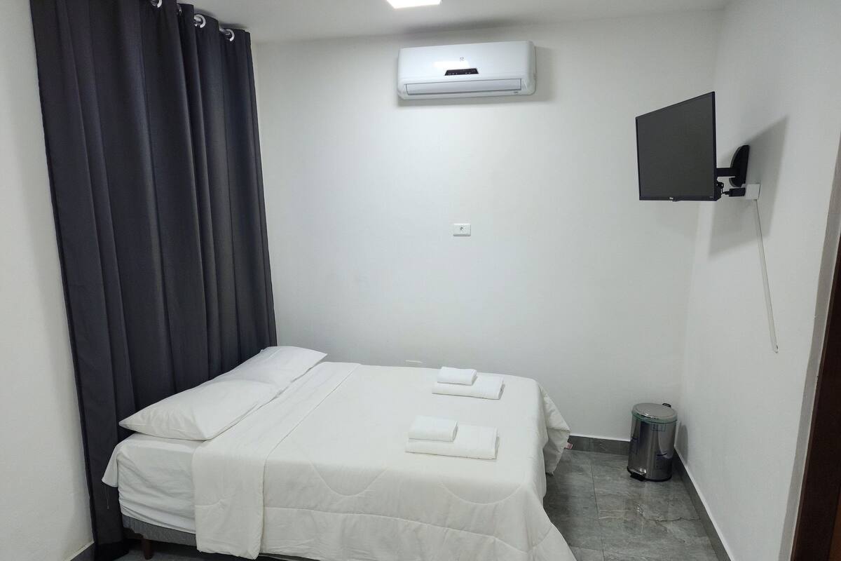 A small, clean room with a bed, a TV mounted on the wall, and an air conditioning unit above the bed.