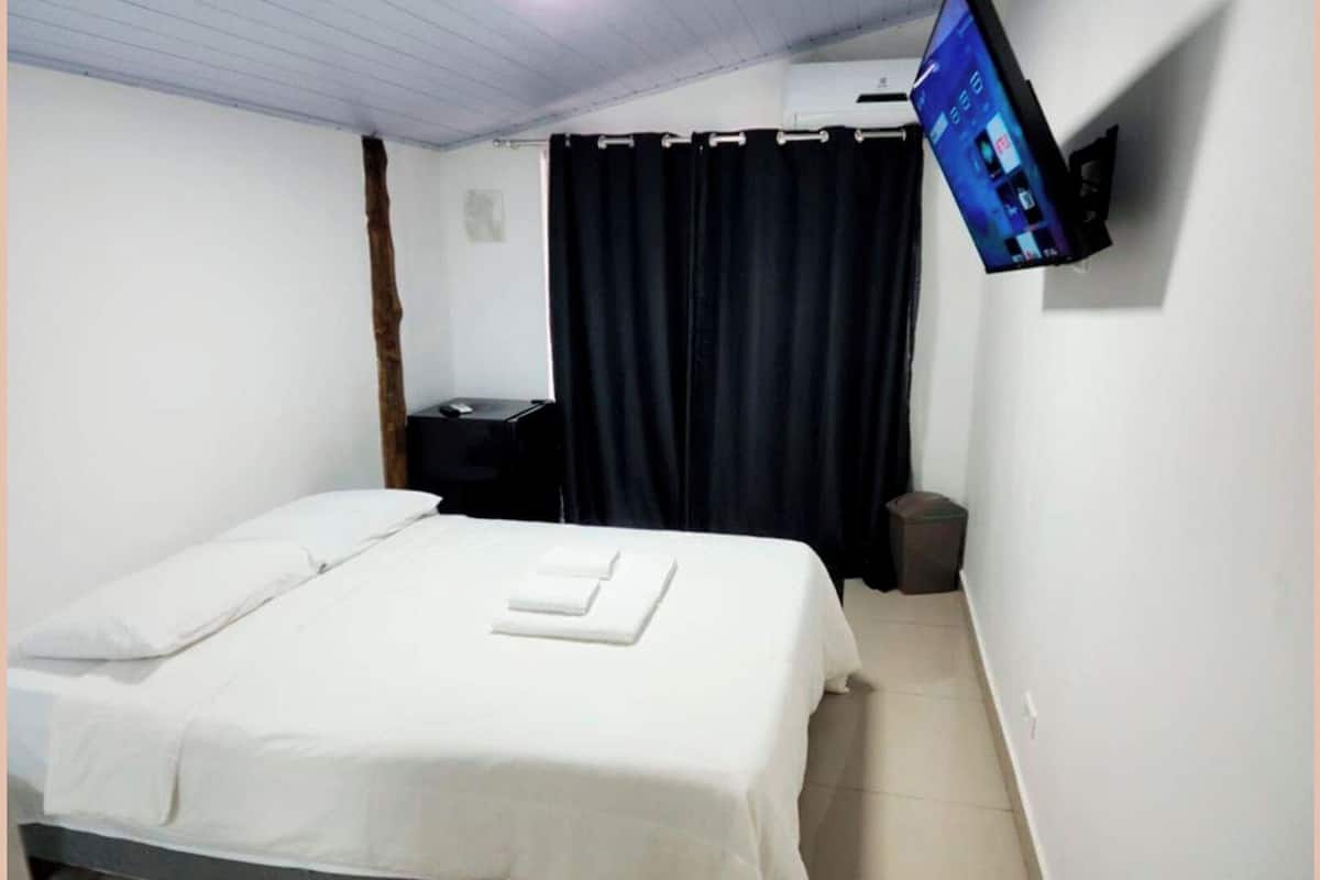 A single bed with white bedding, a flat-screen TV mounted on the wall, a desk with a computer, and a trash bin.