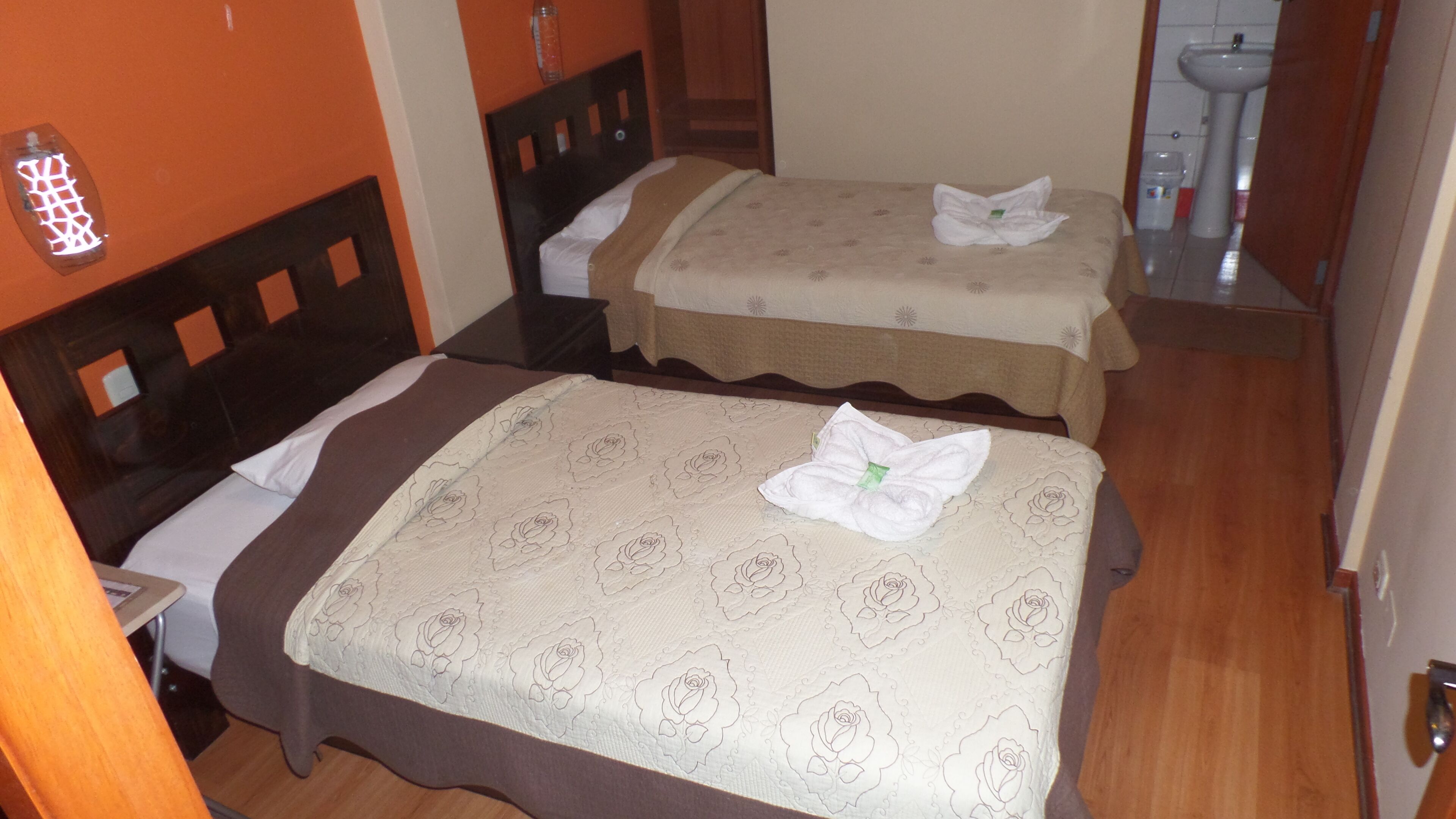 A hotel room with two beds, a wooden headboard, a bedside table, and a bathroom with a sink and mirror.