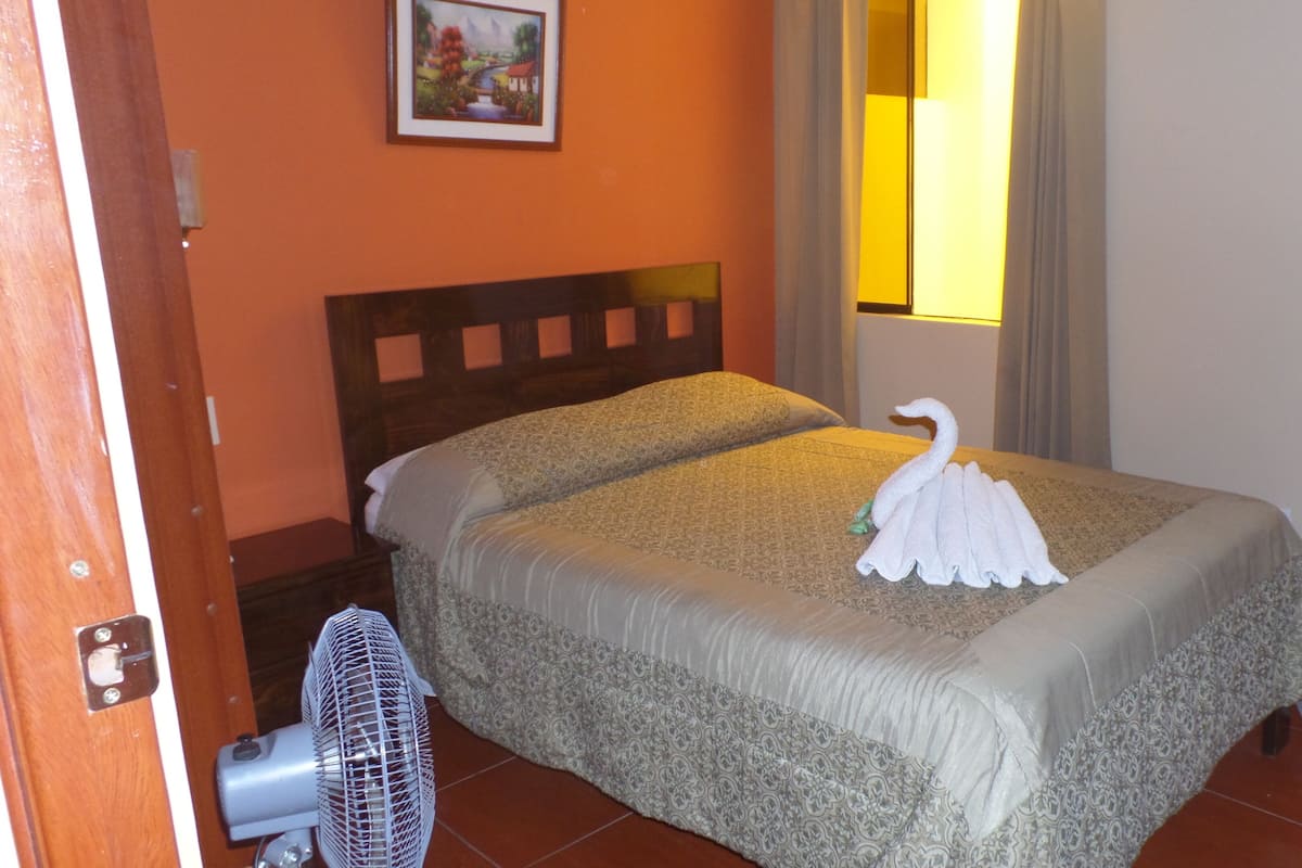 A hotel room with a bed, a fan, and a painting on the wall.