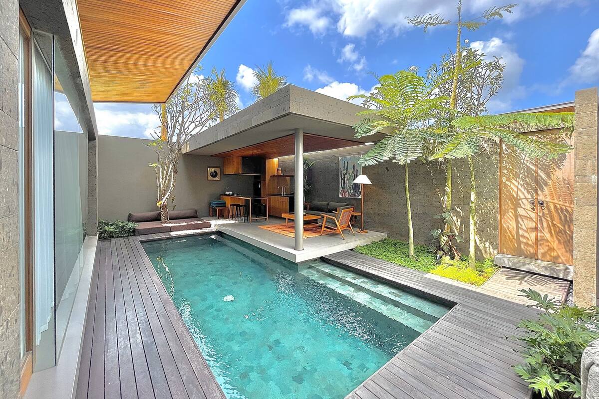 A modern outdoor pool area with a wooden deck, a covered seating area, and a stone wall.