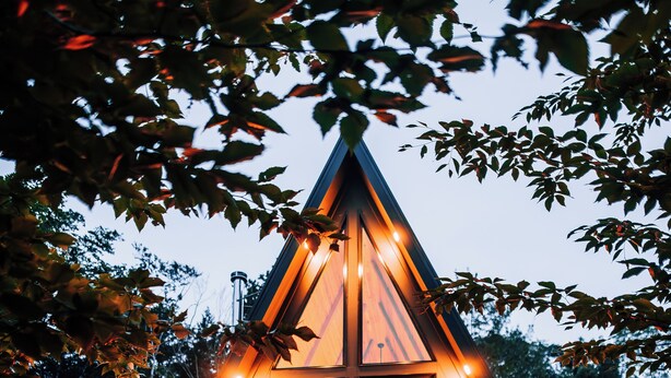 Une cabane en bois de forme triangulaire, dotée de grandes fenêtres et d’un porche, entourée d’arbres.