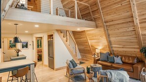 Un loft spacieux avec de hauts plafonds, des poutres apparentes, un coin repas, un salon avec un canapé et des fauteuils, et un escalier menant à un niveau supérieur.