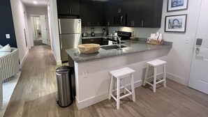 Eine moderne Küche mit dunklen Schränken, einem Edelstahlkühlschrank, einer Granitarbeitsplatte und einer Holznäpfe. Zwei weiße Barhocker und ein Edelstahl-Mülleimer sind ebenfalls vorhanden.