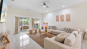 A spacious living room with a large sofa, a coffee table, and a view of the backyard through glass doors. - New*Mini-Golf*Heat or Chill Saltwater Pool*Arcade (Jensen Beach)