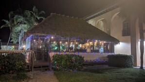 Una estructura de techo de paja con cerramiento de vidrio, rodeada de follaje e iluminada con luces de colores.