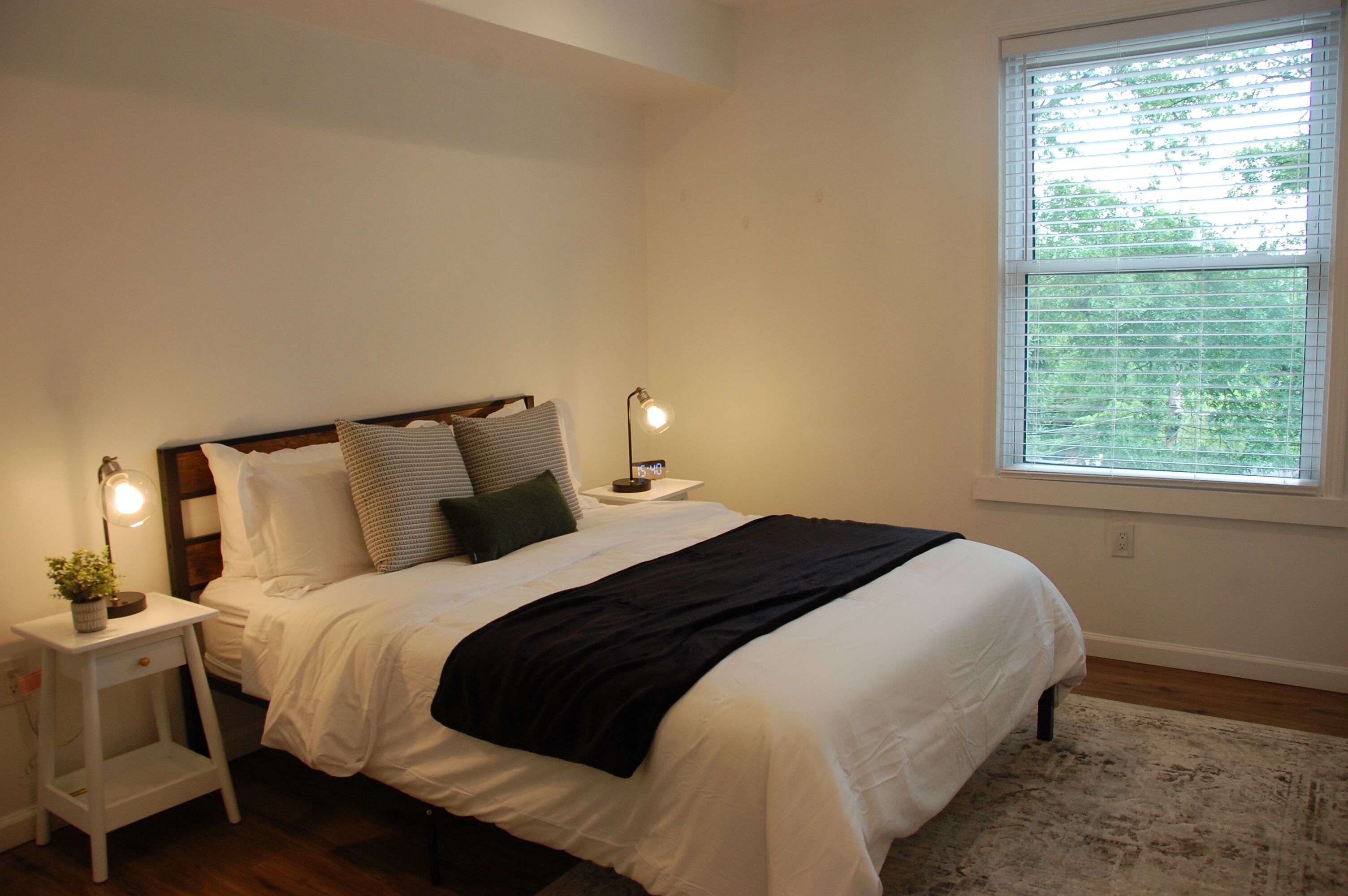 A neatly made bed with white bedding and a black throw, two bedside lamps, a small nightstand with a plant, and a window with blinds.