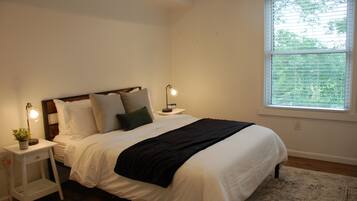 A neatly made bed with white bedding and a black throw, two bedside lamps, a small nightstand with a plant, and a window with blinds.