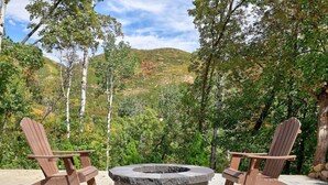 Two Adirondack chairs facing a circular stone fire pit on a concrete patio with a backdrop of trees and hills. - 20% Off Valentines Studio at Sundance Utah (Sundance)