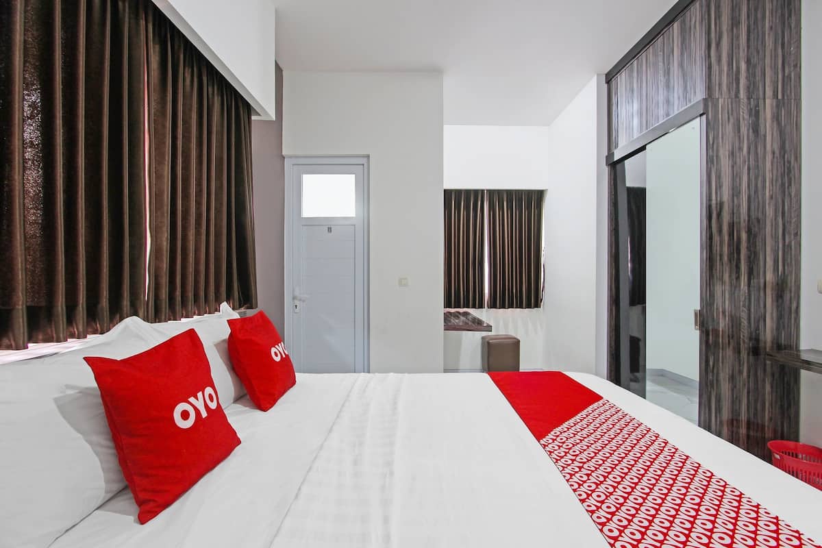 A hotel room with a large bed, two red pillows with "OYO" branding, a red and white patterned runner, a small stool, and a sliding glass door leading to a balcony.