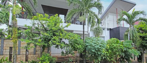 A modern two-story house with a stone facade, a balcony, and lush greenery including palm trees.