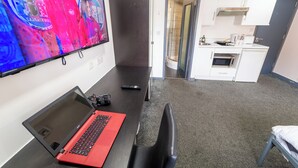 Een kamer met een bureau, een laptop, een stoel, een televisie, een kitchenette en een douche.