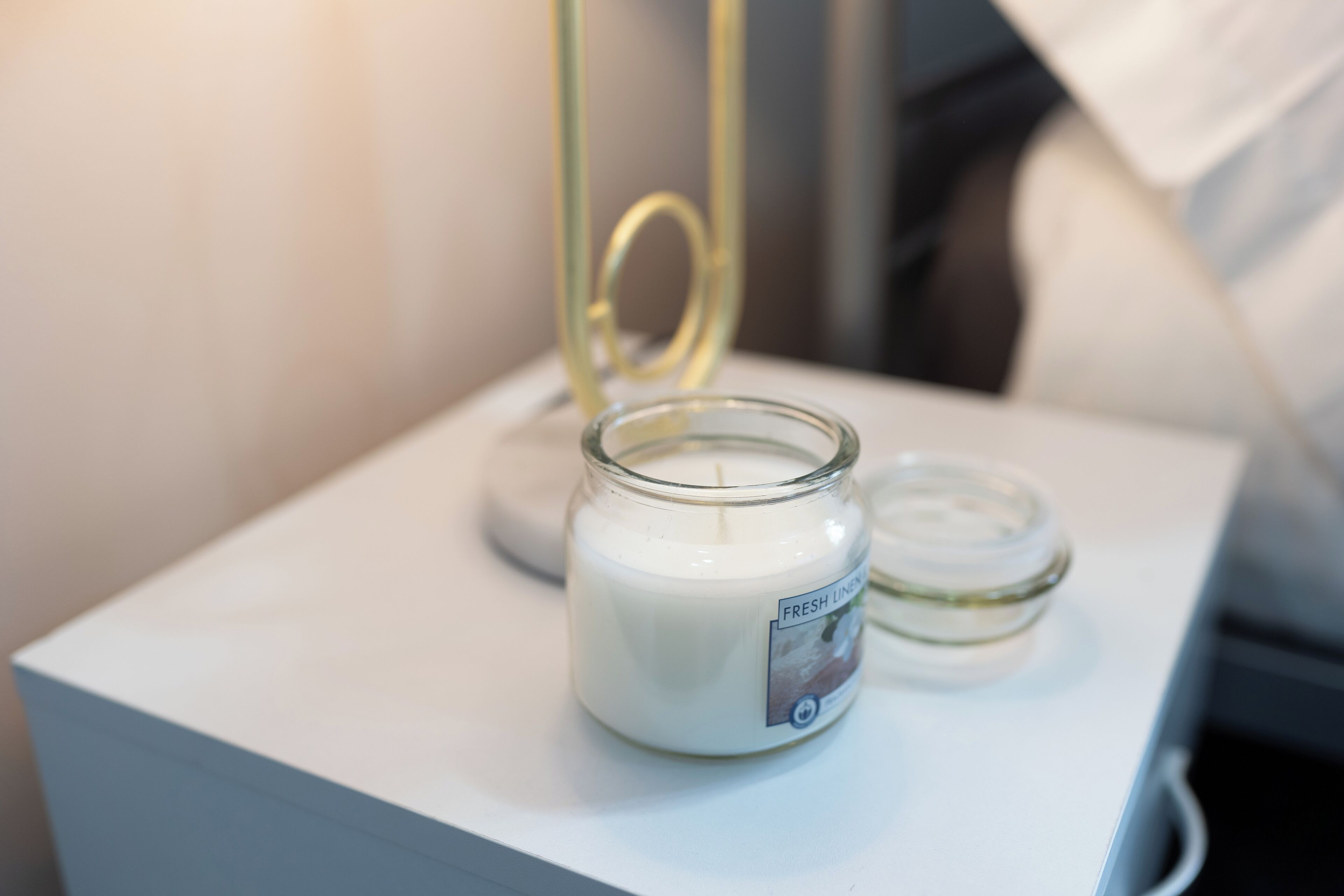 A white bedside table with a glass jar candle labeled "Fresh Linen" and a smaller jar candle beside it.