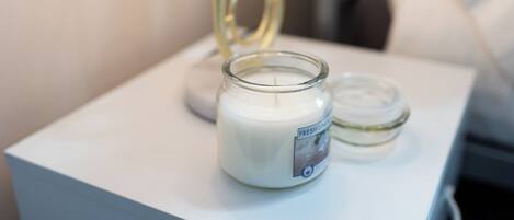 A white bedside table with a glass jar candle labeled "Fresh Linen" and a smaller jar candle beside it.