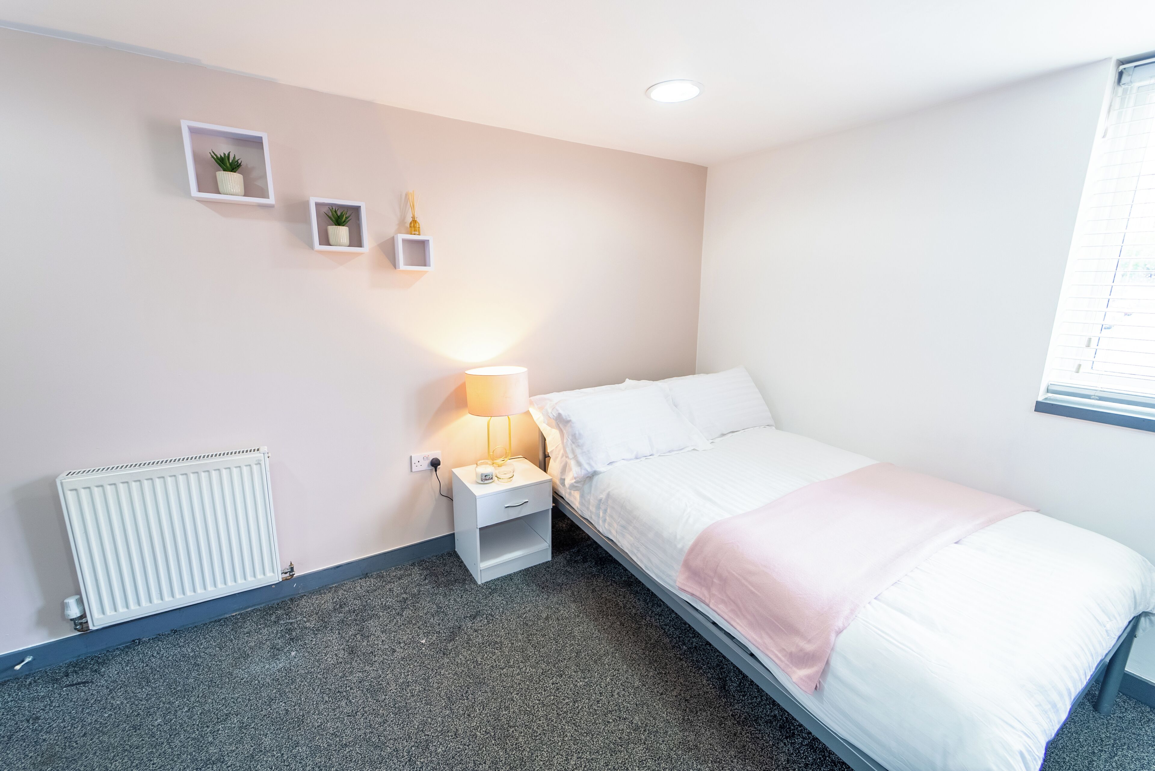 A single-bed room with a white bed, a bedside table with a lamp, a radiator, and two wall-mounted picture frames.