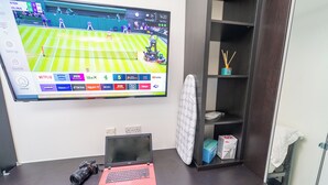 A hotel room with a desk featuring a laptop, a camera, and a flat-screen TV displaying a tennis match.