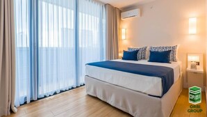 Una habitación de hotel con una cama grande, dos mesitas de noche con lámparas, un equipo de aire acondicionado y cortinas traslúcidas.