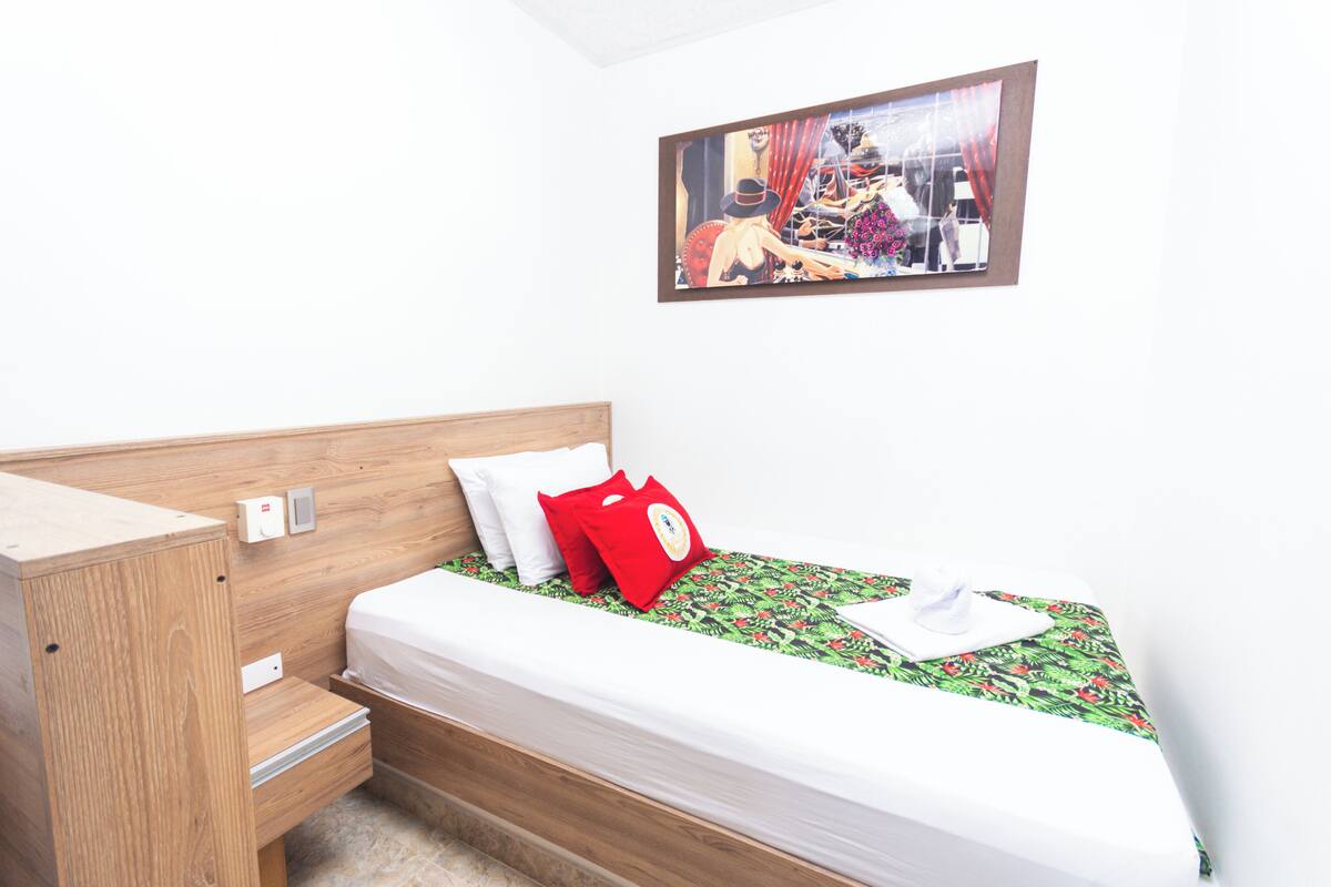 A small, neatly arranged bedroom with a wooden headboard, a single bed with a patterned bedspread, a red pillow, and a wall-mounted painting.