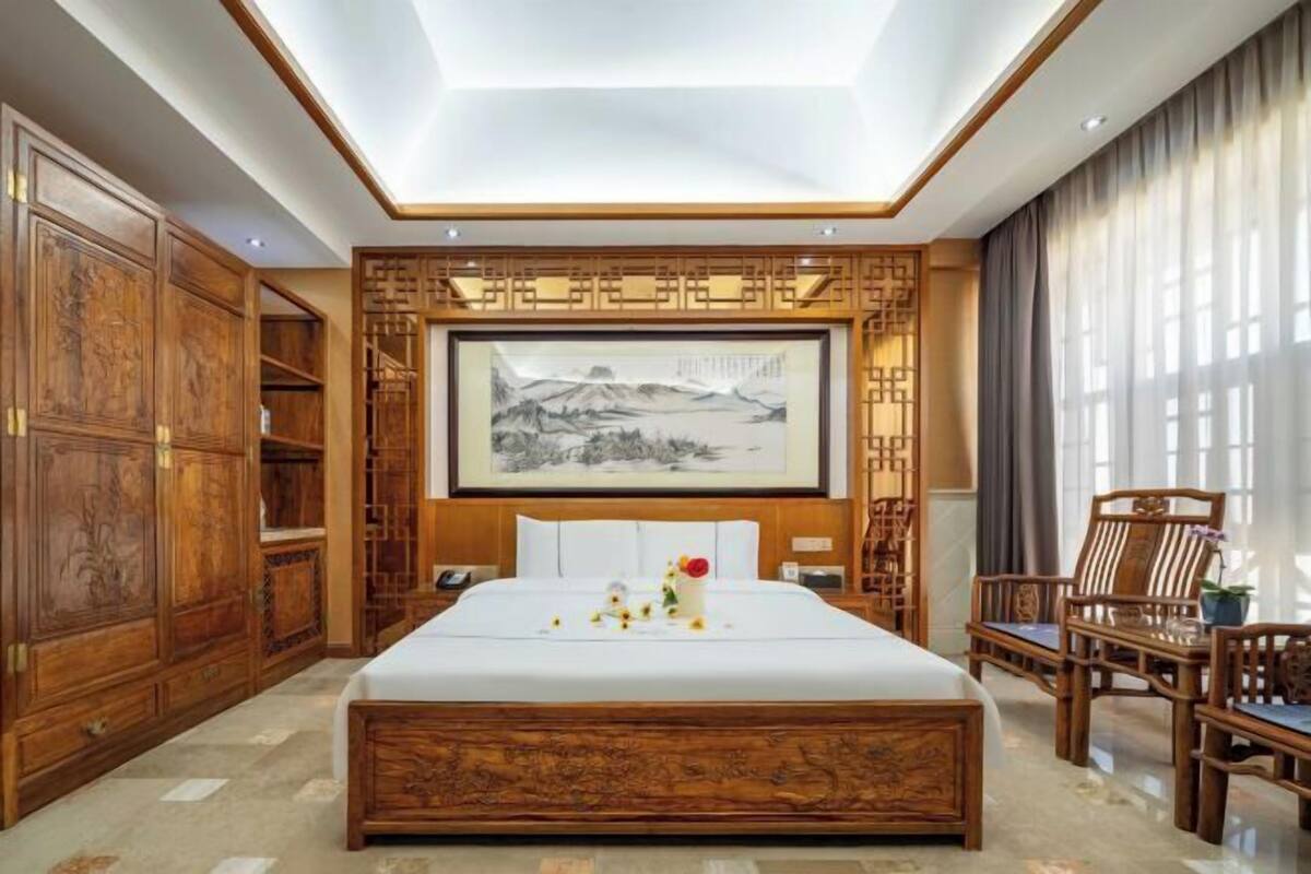 A hotel room with a large bed, wooden furniture, a painting on the wall, and a view of the outdoors.