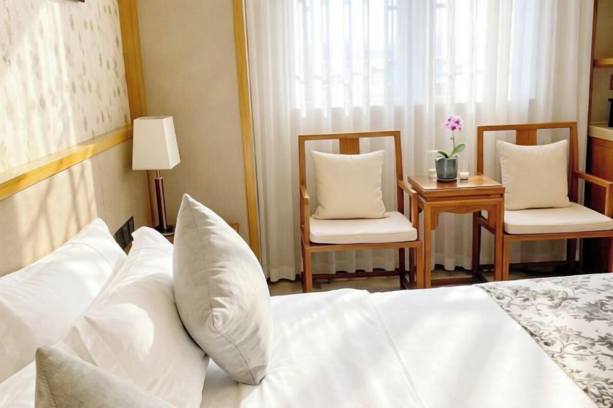 A hotel room with a bed, two chairs, a small table with a vase of flowers, and a lamp.
